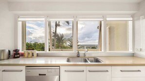 A modern kitchen with a dishwasher, a sink, and a window with a view of palm trees and a cloudy sky. - Picture Perfect - Panoramic Views, Walk to Beach (Noosa Heads)
