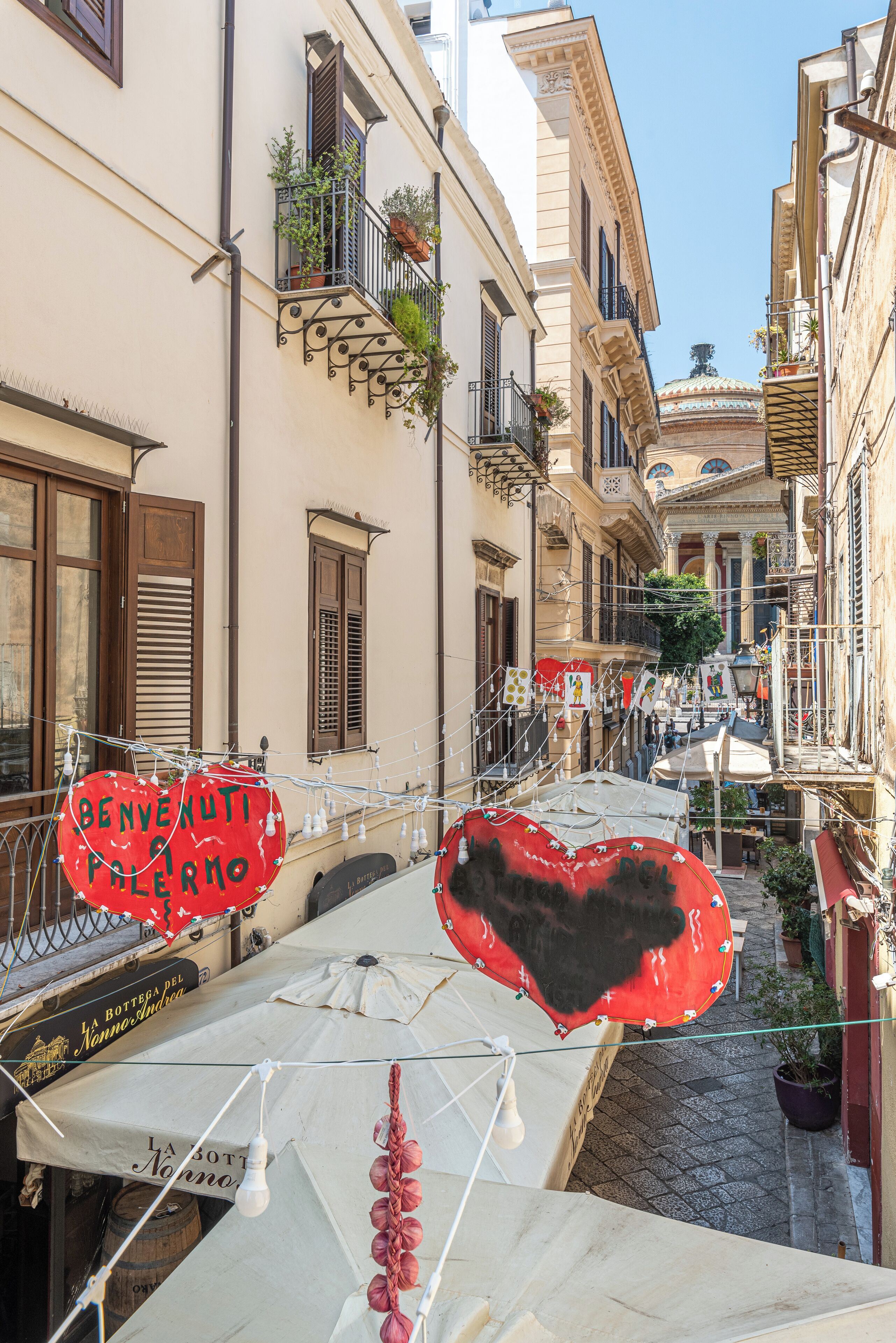 A narrow street with buildings on either side, some with balconies and plants. There are heart-shaped decorations and a sign that reads "BENVENUTI PELLEMO".