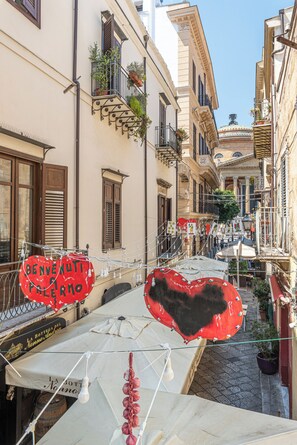 A narrow street with buildings on either side, some with balconies and plants. There are heart-shaped decorations and a sign that reads "BENVENUTI PELLEMO".