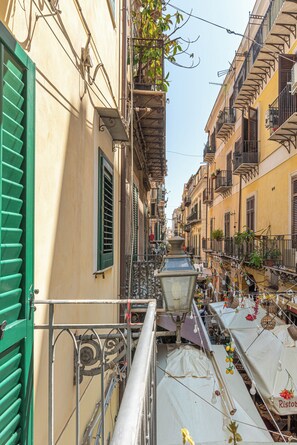 A narrow street with buildings on either side, balconies with plants, and a street lamp.