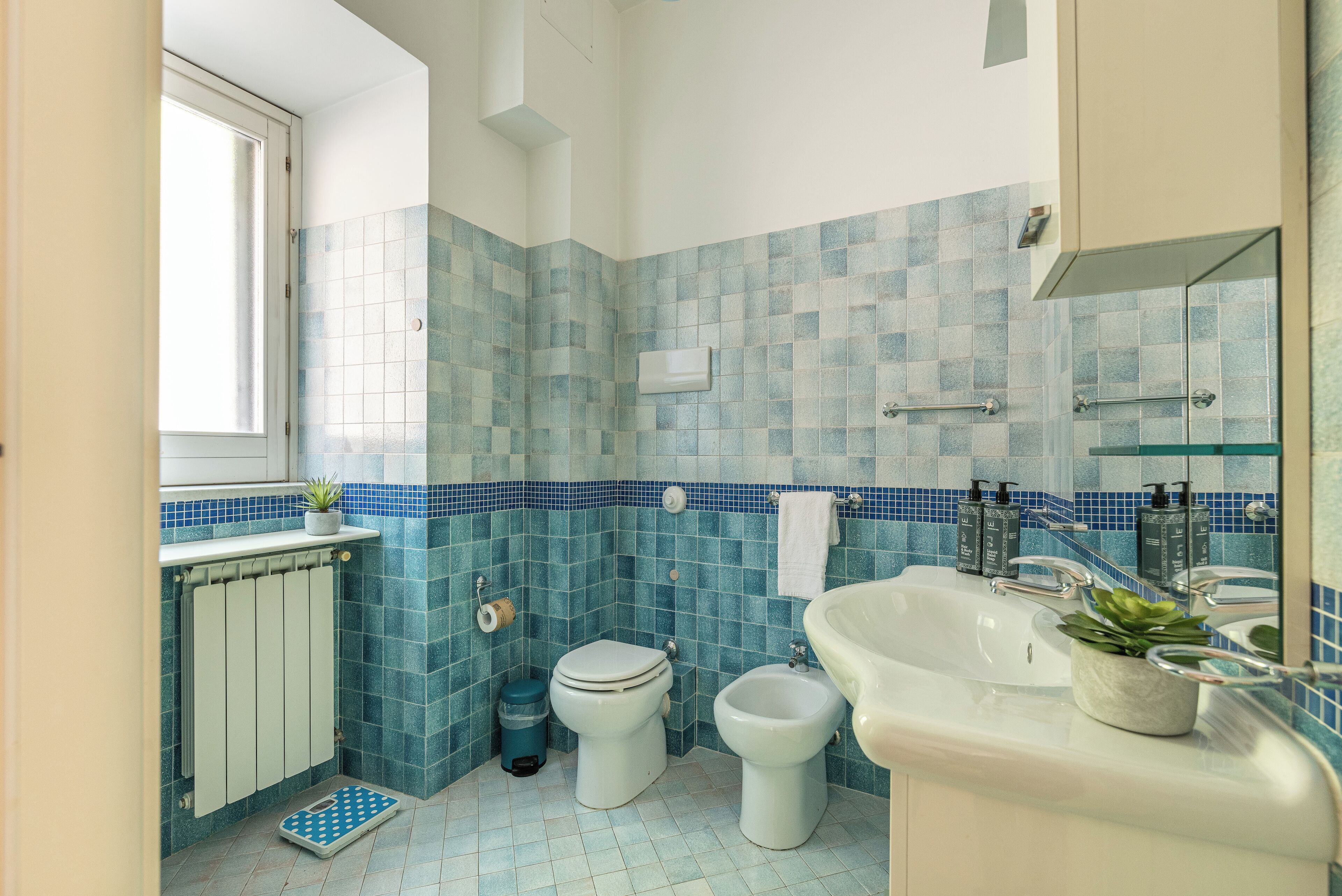 A bathroom with blue mosaic tiles, a white toilet, a bidet, a sink with a mirror, and a window providing natural light.