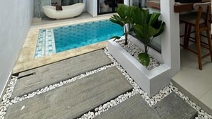 A modern outdoor pool area with a white infinity pool, a wooden deck, and a seating area with a chair and table. - Maithala Villa (Kuta)
