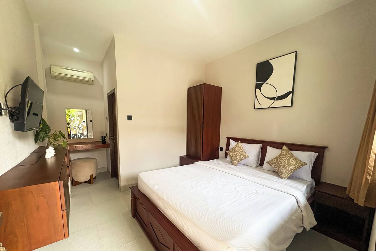 A hotel room with a large bed, a wooden desk, a TV, an air conditioner, and a painting on the wall.