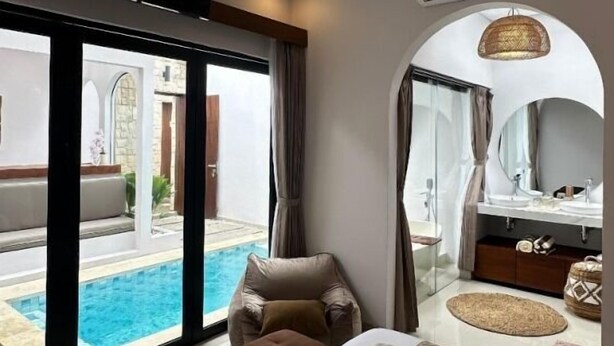 A modern hotel room with a large bed, a sitting area with a view of a pool, a bathroom with a vanity and mirror, and an air conditioning unit on the wall. - Maithala Villa (Kuta)