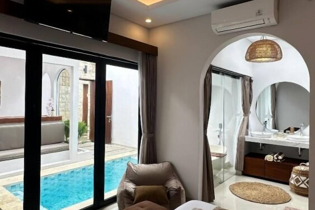 A modern hotel room with a large bed, a sitting area with a view of a pool, a bathroom with a vanity and mirror, and an air conditioning unit on the wall.