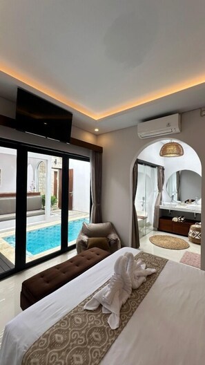 A modern hotel room with a large bed, a sitting area with a view of a pool, a bathroom with a vanity and mirror, and an air conditioning unit on the wall. - Maithala Villa (Kuta)