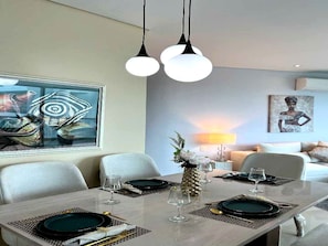 A dining area with a long table set for two, two pendant lights, a painting on the wall, and a sofa in the background. - Luxurious 3-bedroom Apartment With Shared Pool in Bolívar (Cartagena)