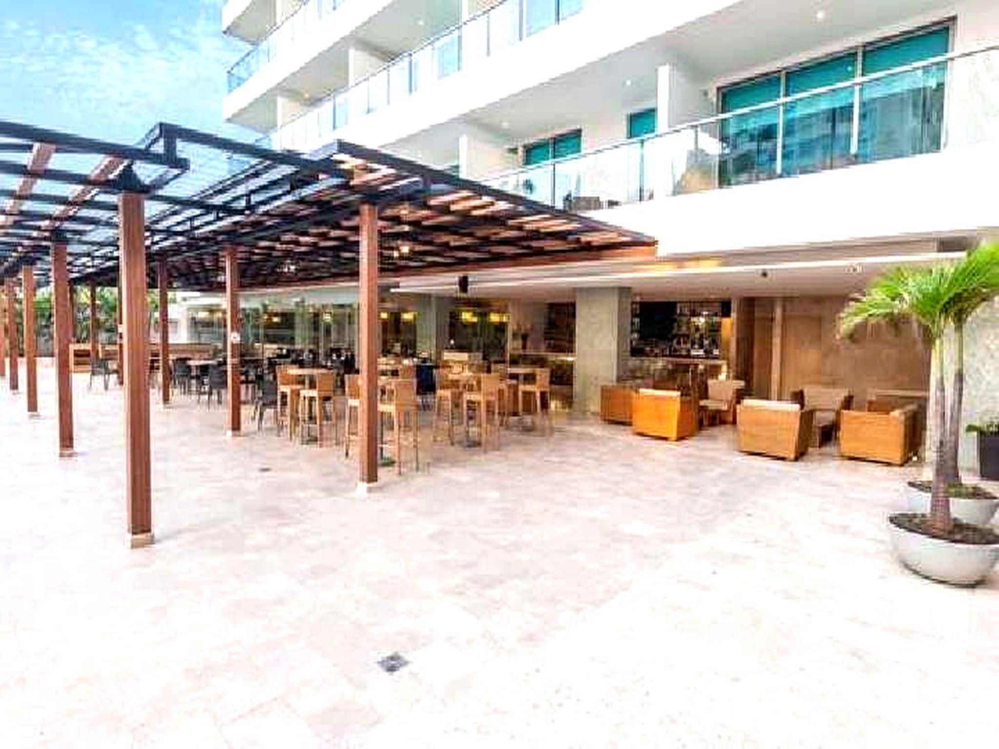 Un espacio al aire libre cubierto con mesas y sillas, una zona de bar con asientos, y una planta grande en maceta.