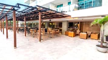 Un espacio al aire libre cubierto con mesas y sillas, una zona de bar con asientos, y una planta grande en maceta.