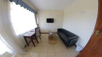 Un petit salon avec un canapé, un téléviseur fixé au mur, une table à manger avec des chaises et une fenêtre avec des rideaux.