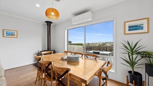 Un coin repas avec une table en bois, des chaises, un luminaire suspendu et une grande fenêtre donnant sur la mer.