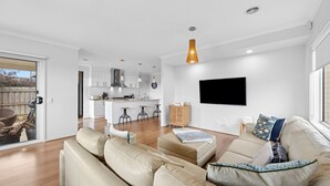 Un salon moderne avec un canapé, un téléviseur fixé au mur et une cuisine en arrière-plan.