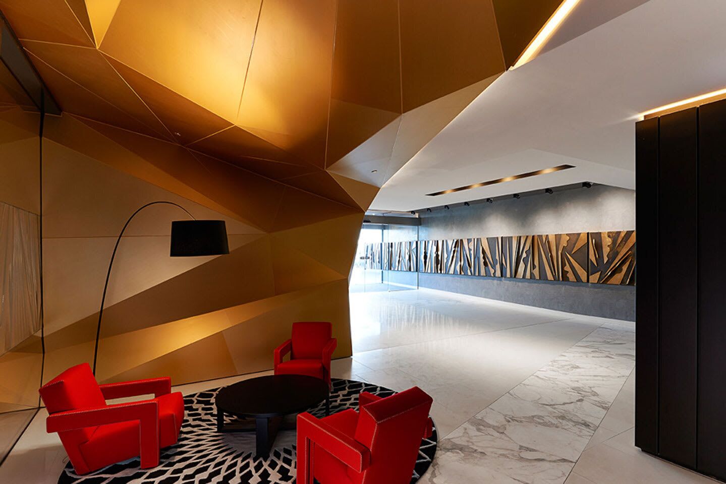 A modern lobby with geometric ceiling design, red seating, and a patterned wall.