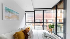 Une chambre à coucher moderne avec une grande fenêtre, un lit recouvert d’une literie blanche à motifs et une œuvre d’art encadrée accrochée au mur.