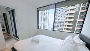 Habitación de hotel moderna con una cama grande, ropa de cama blanca, vistas de edificios altos y una puerta corrediza que da a un balcón.