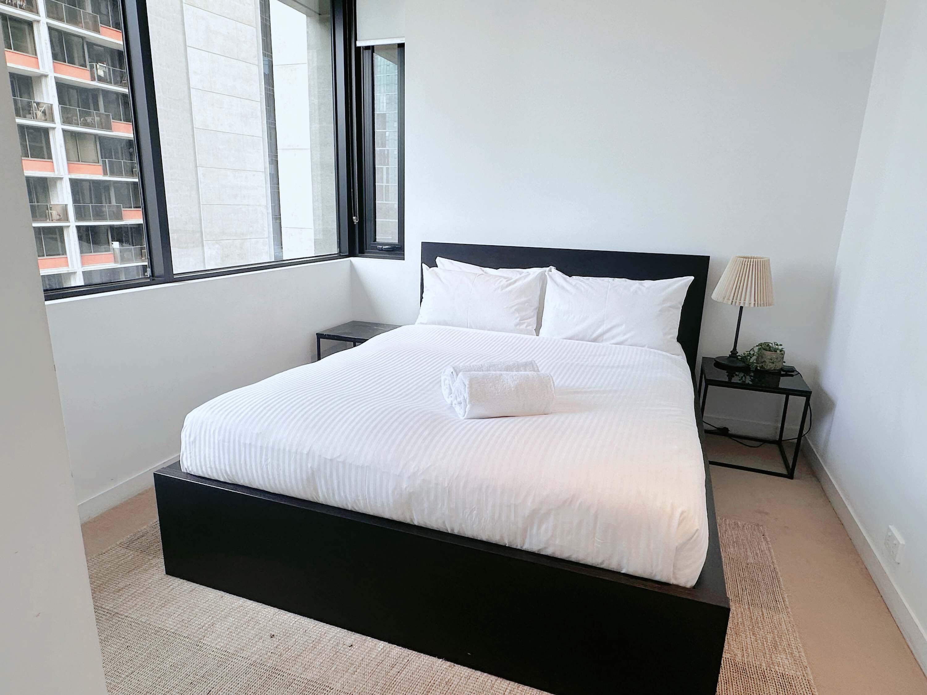 A modern bedroom with a black bed frame, white bedding, a small bedside table with a lamp, and a view of a building through a large window.