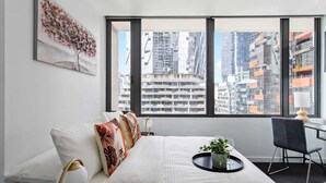 A modern hotel room with a large window, a bed with white bedding, a side table with a plant and glasses, and a city view. - A Cozy 2BR Apt Next to Southern Cross Free Parking (Melbourne)