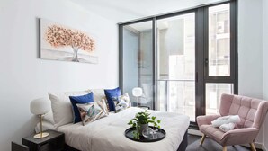 A modern bedroom with a large bed, a side table, a chair, a window with a view, and a painting on the wall. - A Cozy 2BR Apt Next to Southern Cross Free Parking (Melbourne)