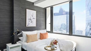 Ein modernes Hotelzimmer mit einem großen Bett, einem Nachttisch mit Lampe, Blick auf eine Stadt durch große Fenster und einem gerahmten Kunstwerk an der Wand.