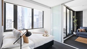 Ein modernes Schlafzimmer mit einem großen Bett, einem Schreibtisch und einem Stuhl. Große Fenster bieten Blick auf die Stadt, eine Glastür führt in einen Wohnbereich und eine Zimmerpflanze sorgt für Frische.