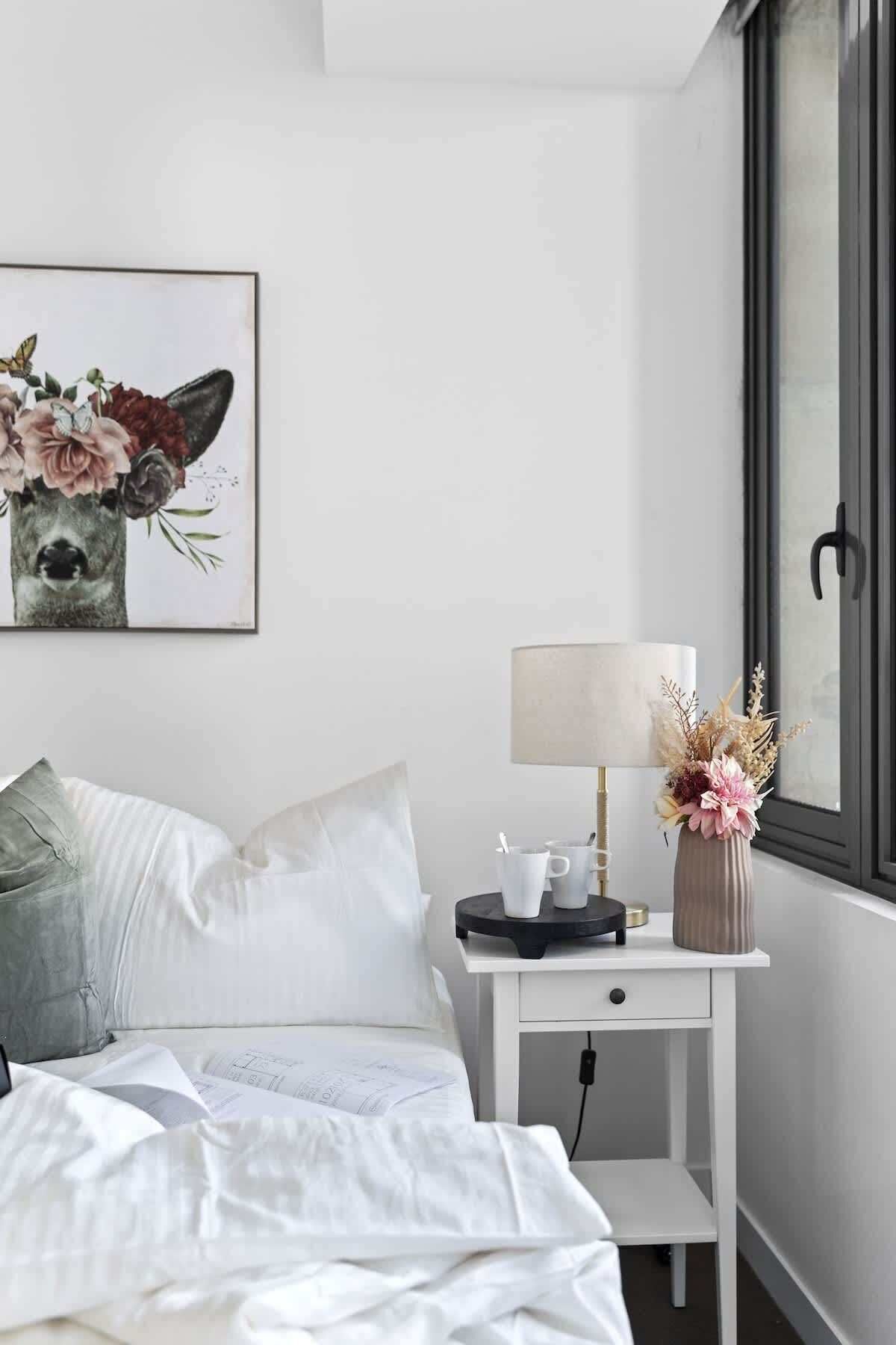 A neatly made bed with a pillow, a bedside table with a lamp and a vase of flowers, and a framed picture on the wall.