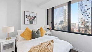 Une chambre d’hôtel moderne avec un grand lit, une table de chevet, une lampe, une œuvre d’art florale encadrée et une vue sur les bâtiments de la ville, visible à travers une grande fenêtre.