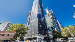 A modern high-rise building with a prominent glass facade, surrounded by other tall buildings and a busy street scene. - A Cozy & Modern 2BR Apartment, Fantastic Views (Melbourne)