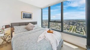 A bedroom with a large window, a bed, a bedside table with a lamp, and a framed picture on the wall. - A Cozy & Modern 2BR Apartment, Fantastic Views (Melbourne)