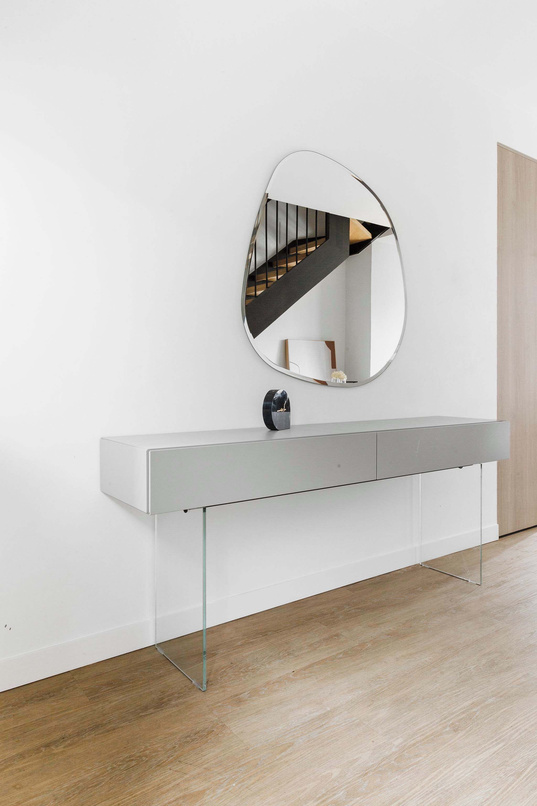 A modern, minimalist room with a white console table, a large oval mirror, and a wooden staircase in the background.