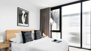Moderni hotellihuone, jossa on suuri sänky, yöpöytä, kehystetty taideteos ja näkymä suuresta ikkunasta.