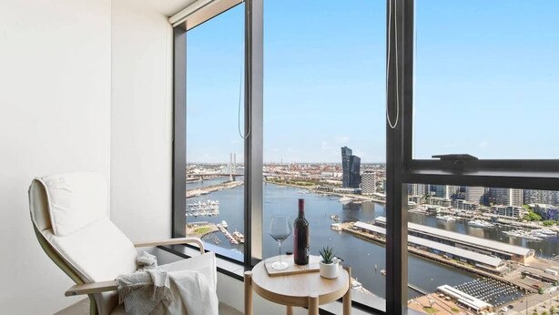 A balcony with a view of a river and cityscape, a chair, a small table with a wine bottle and glass, and a plant.
