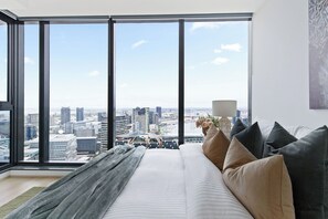 A high-rise hotel room with a large window offering a city view, a bed with multiple pillows, and a lamp on the nightstand. - A Cozy 2BR Apt With Harbour Views, Free Parking (Melbourne)