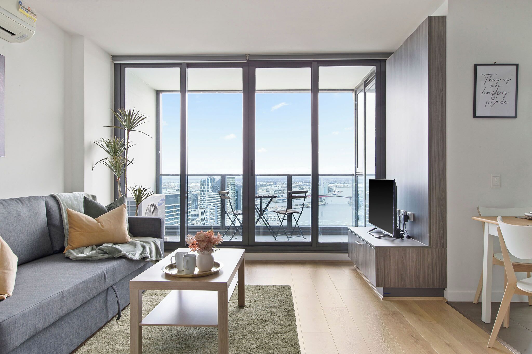 A modern living room with a grey sofa, a coffee table, and a TV on a wooden stand. There is a balcony with a view of the city.