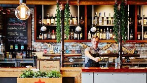 Un bar avec un tableau noir affichant la carte des boissons, un barman en train de travailler et un comptoir en bois sur lequel se trouve une caisse de bière.