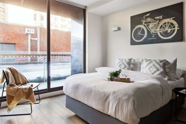 A modern bedroom with a large bed, a view of the street through a sliding door, and a motorcycle-themed artwork on the wall.