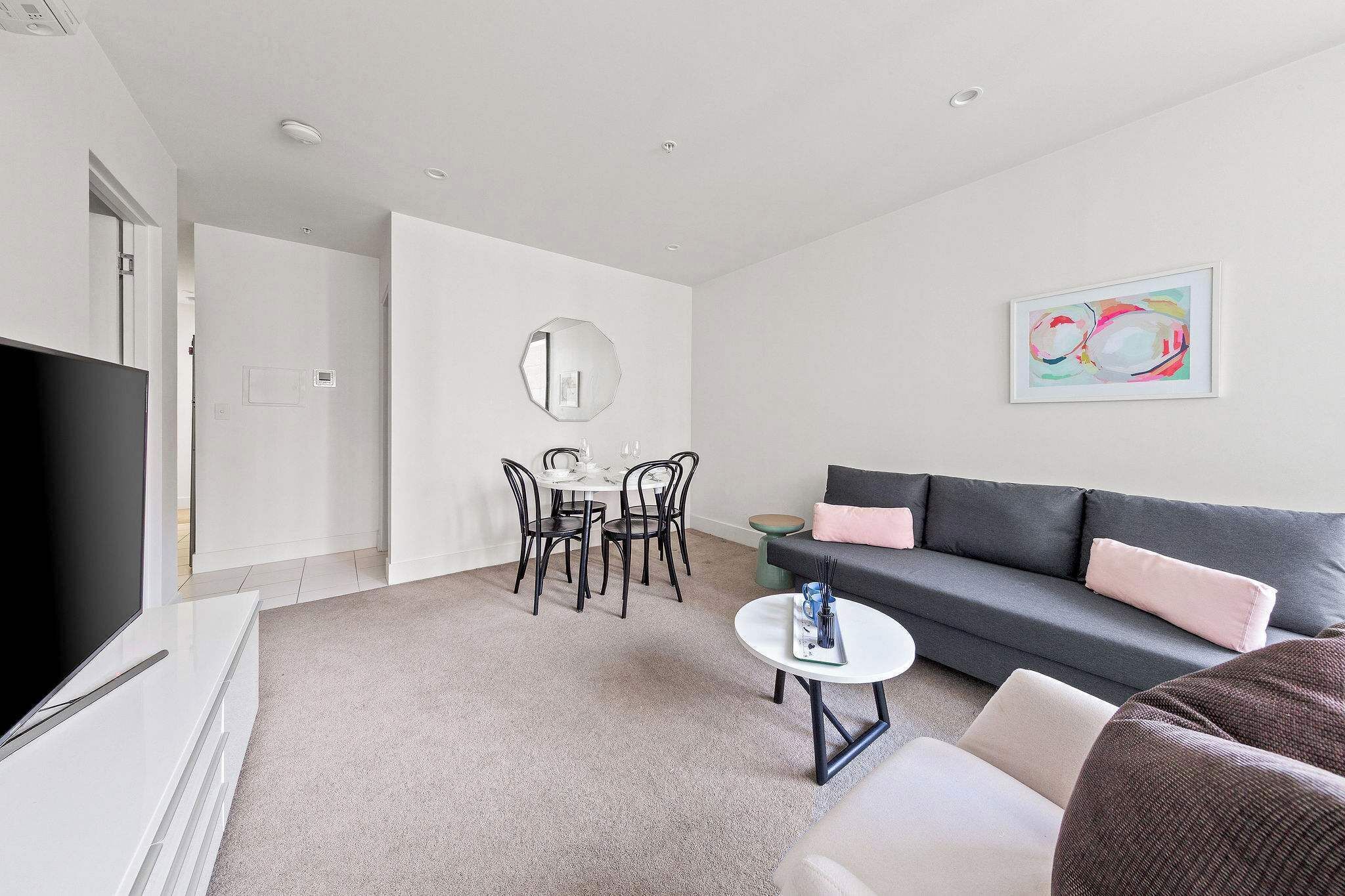En moderne stue med et fladskærms-tv, en grå sofa, et rundt sofabord og et spiseområde med et hvidt bord og sorte stole.