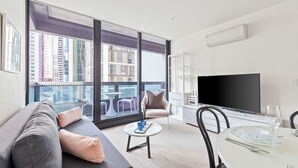 A modern living room with a sofa, a dining table set for two, a flat-screen TV, and a balcony with a view of high-rise buildings. - City View 2br, Free Parking, Bright & Central Apt (Melbourne)