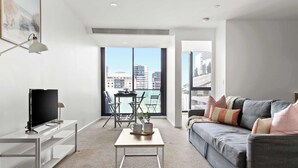 Un salon moderne avec un canapé gris, un téléviseur à écran plat posé sur un meuble blanc, une petite table basse blanche et un balcon offrant une vue sur les immebles.