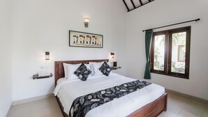 A bedroom with a wooden bed, white bedding, a decorative pillow, a wall-mounted light, a window with curtains, and a small shelf with a framed picture. - Sundhara View Ubud (Sukawati)