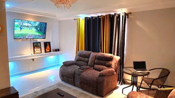 Een moderne woonkamer met een grote flatscreen-tv, een bruine bank, een klein rond tafeltje met een laptop en een kroonluchter.