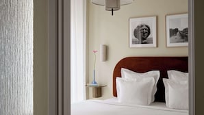A hotel room with a bed, white pillows, a nightstand with a vase and books, and two framed pictures on the wall.