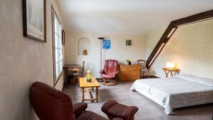 Una habitación con una cama, dos sillones de cuero marrón, una mesa de madera con un jarrón, una silla roja, un aparador y una lámpara.