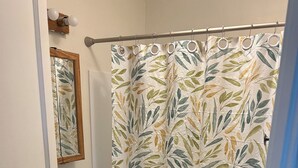 Un baño con lavamanos blanco, espejo, cortina de ducha con estampado de hojas y alfombra de baño negra.