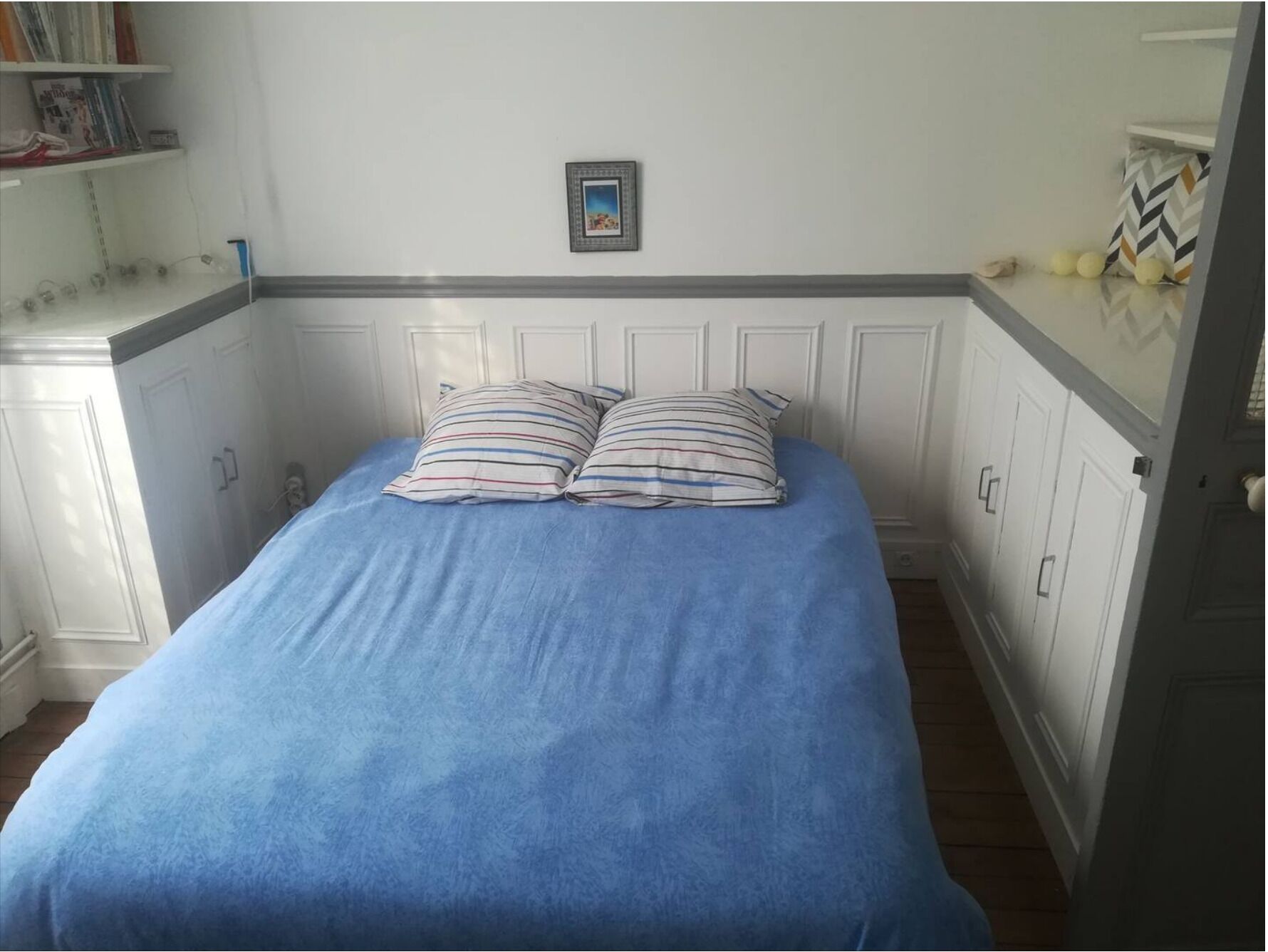A bedroom with a bed, two pillows, a picture on the wall, and a shelf with books and decorative items.