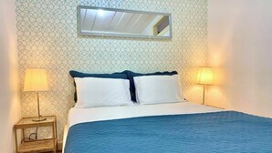 A neatly made bed with a blue quilt, two white pillows, and a wooden nightstand with a lamp. - Central Avenue #1 (Lisboa)