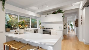Eine moderne Küche mit weißen Schränken, einer großen Kochinsel mit Barhocker und Blick ins Freie durch große Fenster.