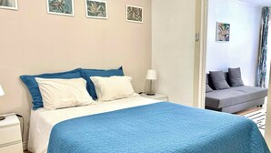 A neatly made bed with a blue quilt, a bedside table with a lamp, and a grey sofa in the background. - Central Avenue #2 (Lisboa)