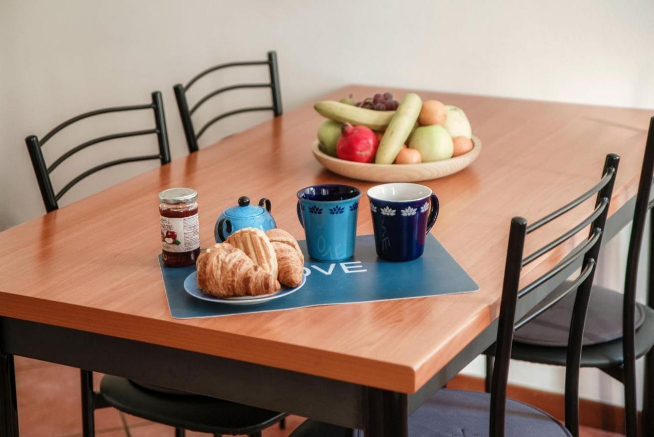Un tavolo da pranzo in legno con una ciotola di frutta, un vasetto di marmellata, un croissant e due tazze blu decorate con motivi floreali.