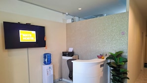 A reception area with a flat-screen TV displaying "MAYS HOSTEL" sign, a water dispenser, a printer, a desk, and a potted plant. - Mays Hostel (Hua Hin)