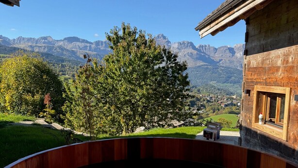 A wooden hot tub with a view of mountains and a building in the background. - Chalet “Mazot De La Pointe Percée” with Balcony (Combloux)